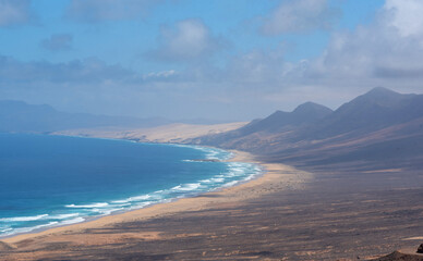 Fototapeta na wymiar Vista panorámica y aérea de la playa de arena blanca y mar cristalino de Cofete en Fuerteventura, rodeada de montañas volcánicas en un día soleado con cielo azul claro en las Islas Canarias.