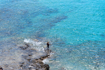 Hombre de espaldas sobre una roca junto al mar pescando con una caña de pescar en las aguas turquesas del mar frente a Fuerteventura en las Islas Canarias en un día soleado de verano con cielo azul