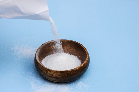 Sugar Is Poured From A White Paper Bag Into A Wooden Bowl On A Blue Background