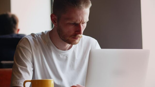 A serious man is typing on the keyboard of his laptop while sitting in a cafe. A man remotely works at a laptop while sitting in a cafe with a cup of coffee.