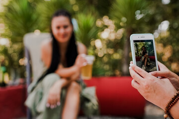 Close-up of a hand taking a photo of a smiling woman with a drink using a smartphone. Summer outdoor scene with palm trees, relaxation, and casual social moments captured digitally.