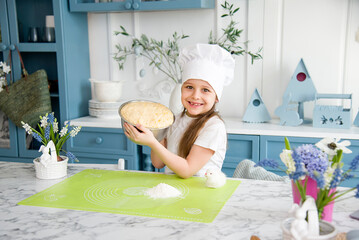 little cute  smiling girl wearing a white chef's hat and uniform holding a bowl of dough for easter bread in the blue and white kitchen