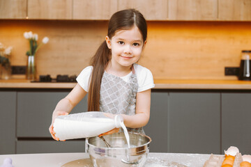 Portrait of a cute little girl standing in a modern kitchen and preparing dough.