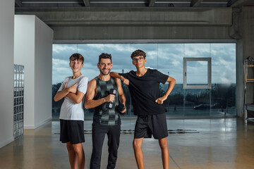 Group of young people resting and looking at camera after training session in gym. Coach and two male teenagers standing at big health club with windows. Healthy lifestyle and friendship concept