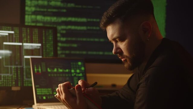 A young man makes notes in a notebook, sitting in front of monitors on which green lines of program code are running. A young hacker writes something in a notebook while a virus program downloads data