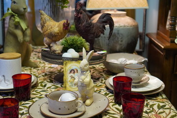 Easter interior of a rabbit figurine with plates on the table and figurines of a chicken and a cockerel on a gray wall background © Irina