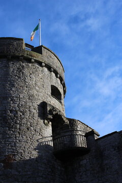 Irish flag atop old castle tower shining in the sun with blue copy space background + clouds. Irish medieval castle travel shot, background, wallpaper, screensaver with text space - King John's Castle