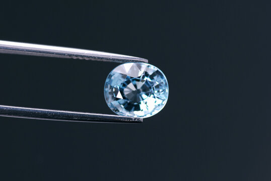 Natural ligh blue aquamarine gemstone in tweezers on dark gray background. Genuine unheated clean transparent semiprecious oval faceted gem setting for making jewelry. Mined in Brazil. Sea blue colour