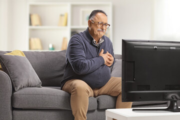 Mature man sitting on a couch and holding his heart in front of tv