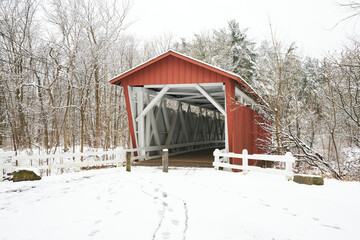 The everett road covered bridge in Cuyahoga Valley National Park. The bridge is red with a white interior. It is a snowy day, and the nearby trees in the forest are covered with fluffy white.