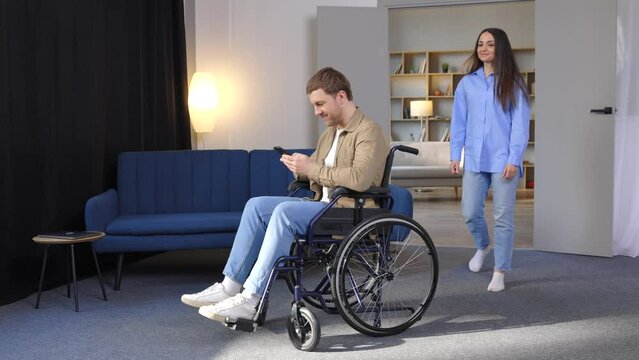 A cute young man in a wheelchair is holding a mobile phone while his girlfriend helps him move around the apartment