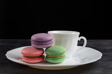 There are macarons on the table in a plate. Behind is a white cup. On a black background. Copy space.