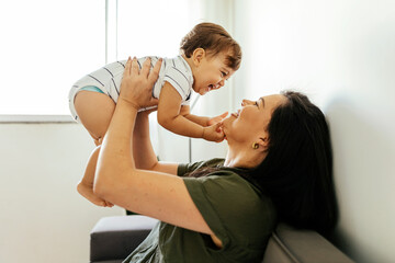 Happy young mother enjoying time with her adorable toddler son at home, lifting him in the air, playing with kid boy in living room