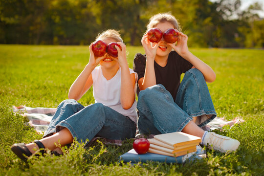 two girls have fun with apples on green grass in park. break in school. girl close eyes with red fresh apples