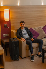 A man with a suitcase is sitting on a sofa in a hotel room while traveling on a business trip