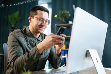 Photo of smiling adult man 30s in green shirt using laptop and mobile phone, while sitting at table...