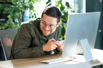 Funny confused office worker happy man employee gamers hold controller joystick playing video game on computer. Male gamer having fun free time at work.