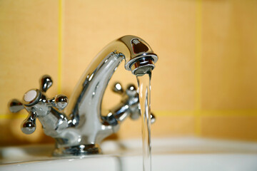 Faucet with calm running water in the bathroom. The focus is on a clean jet against a yellow tile...