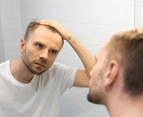 Fototapeta premium Middle aged caucasian white man with a short beard looks at his hair in the mirror in the bathroom and worried about balding. The concept of the problem of male hair loss, early baldness and alopecia