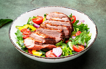 Duck salad with grilled breast and vegetables: red tomato, cucumber, paprika, onoin  and chard, frisse, mizuna and arugula leaves on green table background, top view
