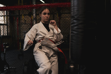 Fototapeta na wymiar A girl in a kimono exercises with a punching bag in the gym while learning karate martial arts