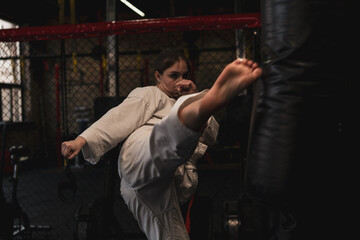 Fototapeta na wymiar A girl in a kimono exercises with a punching bag in the gym while learning karate martial arts