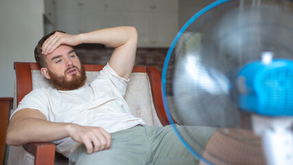 Young bearded man using electric fan at home, sitting on couch cooling off during hot weather,...