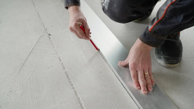 A worker makes markings on the concrete floor with a pencil. Worker using an industrial pencil to measure and mark ceramic tiles at a construction site, preparing flooring material for laying.