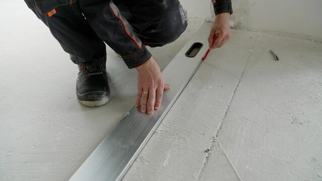 Worker using an industrial pencil to measure and mark ceramic tiles at a construction site, preparing flooring material for laying. A worker makes markings on the concrete floor with a pencil.