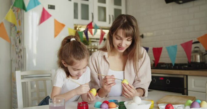 Young mother and daughter painting Easter eggs together at the table in bright modern kitchen. Family gathering in preparation for Easter.