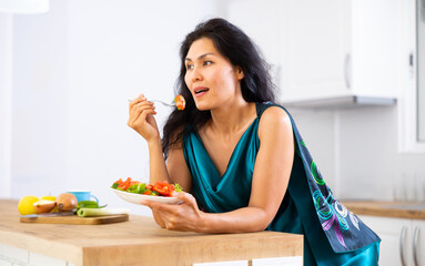 Obraz na płótnie Canvas Asian woman leaning on kitchen table and eating salad. Woman eating vegetable salad in morning, healthy breakfast.