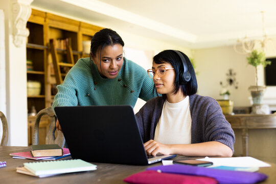 Happy biracial sisters sitting at table and using laptop with headphones - Powered by Adobe
