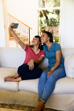 Happy biracial sisters sitting on couch and taking selfie with smartphone