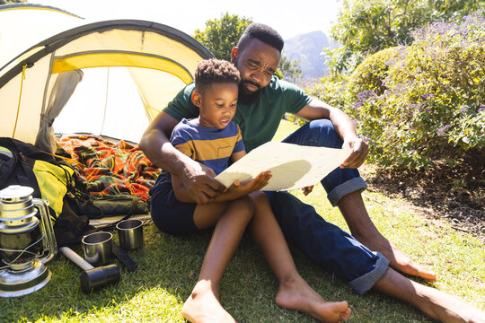 Happy african american father and son sitting on grass and reading map next to tent in garden