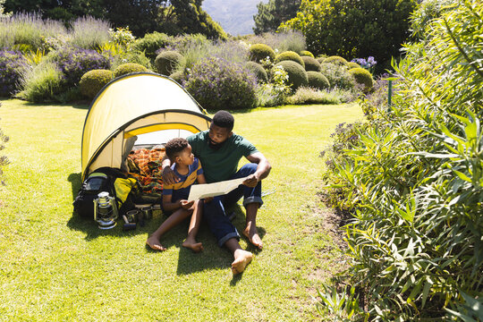 Happy african american father and son sitting on grass and reading map next to tent in garden