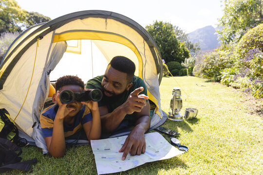 Happy african american father and son lying in tent and looking through binoculars in garden