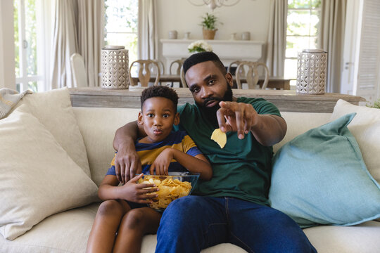 Happy african american father and son sitting on sofa, watching tv and eating crisps