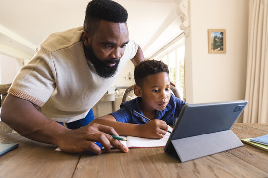 African american father and son sitting at table, doing homework together, using tablet