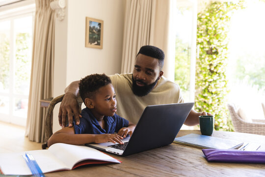 Happy african american father and son sitting at table, doing homework together, using laptop