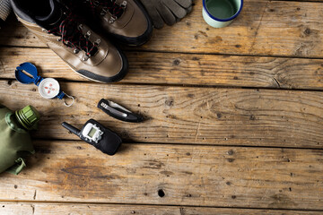 Camping equipment of trekking shoes, metal cup and compass on wooden background with copy space