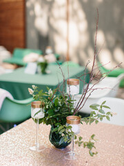 A bouquet of green leaves, white flowers and branches stands in a glass vase. Glass candlesticks on a high leg. Outdoor banquet decor.