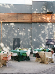 Wedding banquet in green and gold tones in the open air. Bouquets of flowers, colorful chairs on the tables. In the background is a concrete wall with wooden decor.