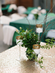 A bouquet of green leaves, white flowers and branches stands in a glass vase. Glass candlesticks on a high leg. Outdoor banquet decor.