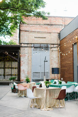 Wedding banquet in green and gold tones in the open air. Bouquets of flowers, colorful chairs on the tables. In the background is a brick building and a large tree.