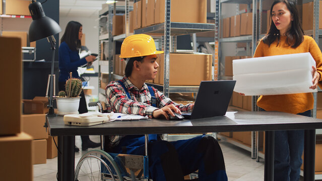Young man in wheelchair looking at stock supplies, checking merchandise in boxes with female worker. Asian adult with impairment planning business development in storage room.