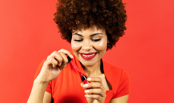 A dark-skinned girl with afro hair posing on a red background with makeup products, the woman is wetting the brush of the liquid lipstick to touch up her lips. Concept of makeup in African people