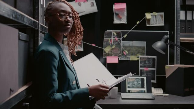African american agent reviewing evidence to solve crime with clues and criminal records, working in police archive. Woman criminologist standing near desk, looking at detective map board.