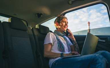 one mature woman caucasian female sitting on the back seat of the car working on laptop computer make a phone call talk in summer day with short gray hair modern on road wearing casual copy space