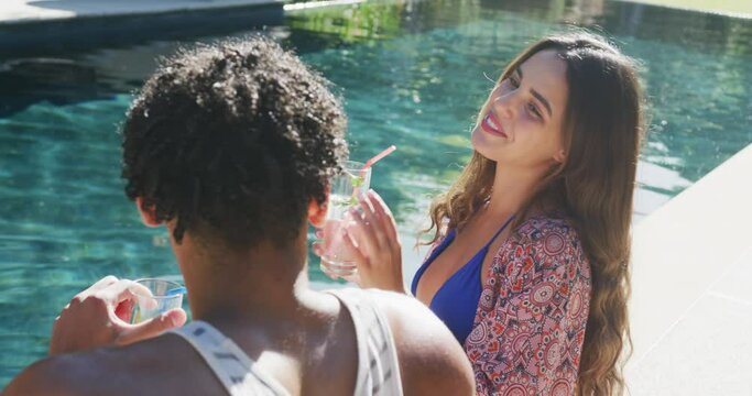 Happy biracial couple talking with drinks at pool in garden on sunny day