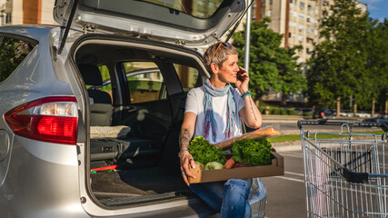 One woman mature caucasian female sit in the back trunk of her car on the parking lot of the supermarket shopping mall or grocery store with vegetables food in box putting them in the vehicle